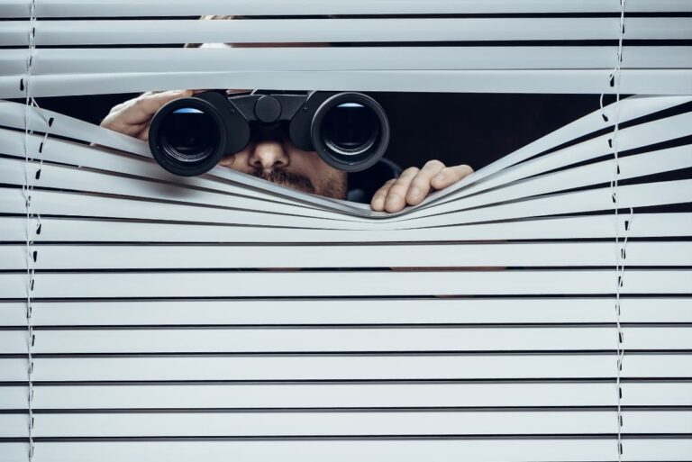 A man with binoculars peeking through the blinds