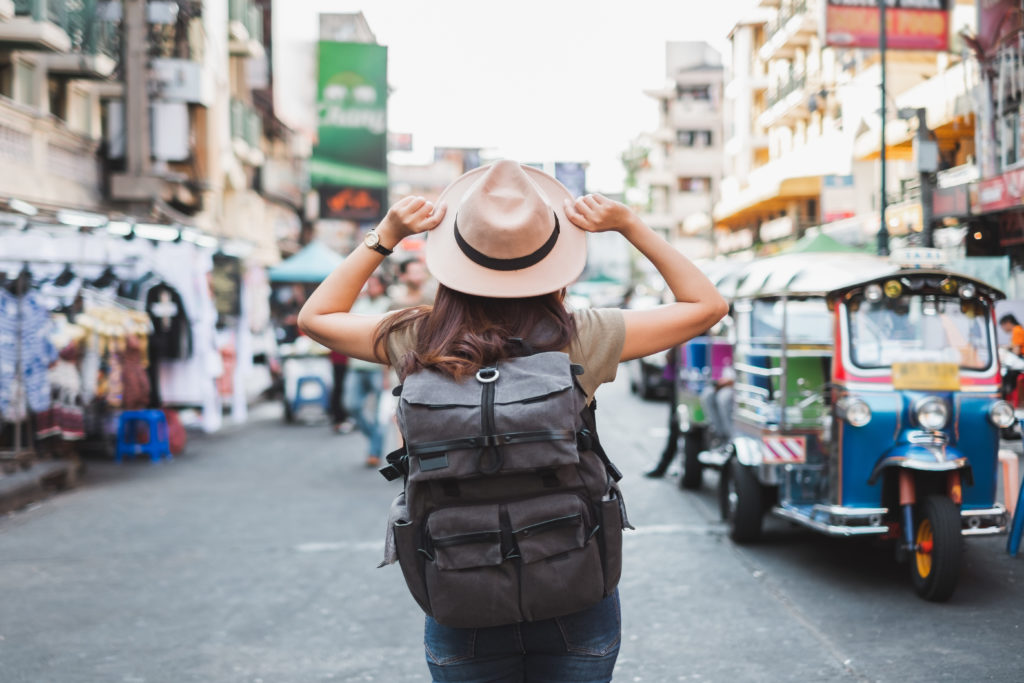 A solo woman traveling internationally. She wears a hat and backpack and stands on a street in Thailand.