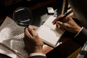 A man in a suit holds a magnifying glass over several pages covered in a cypher and writes in a notebook