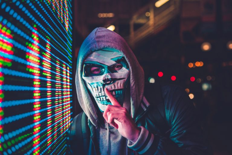 Figure in gray hood and a painted skull mask making “shh” gesture with his forefinger