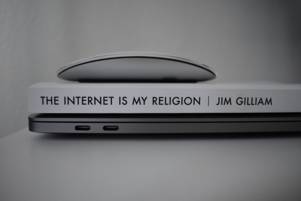A computer mouse sits atop a book that sits atop a laptop. The book’s spine reads The Internet is My Religion.
