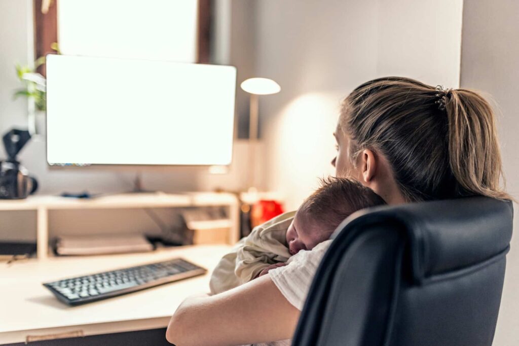 A rear view shows a mother sitting in a chair, holding a baby against her shoulder, as she looks at a computer monitor on a desk.