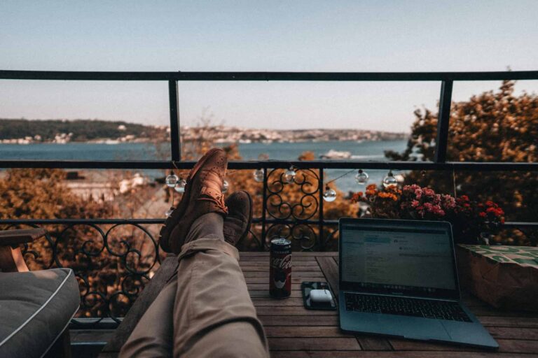 A person relaxing on a balcony beside an open laptop on a table, enjoying a peaceful setting with a panoramic view.