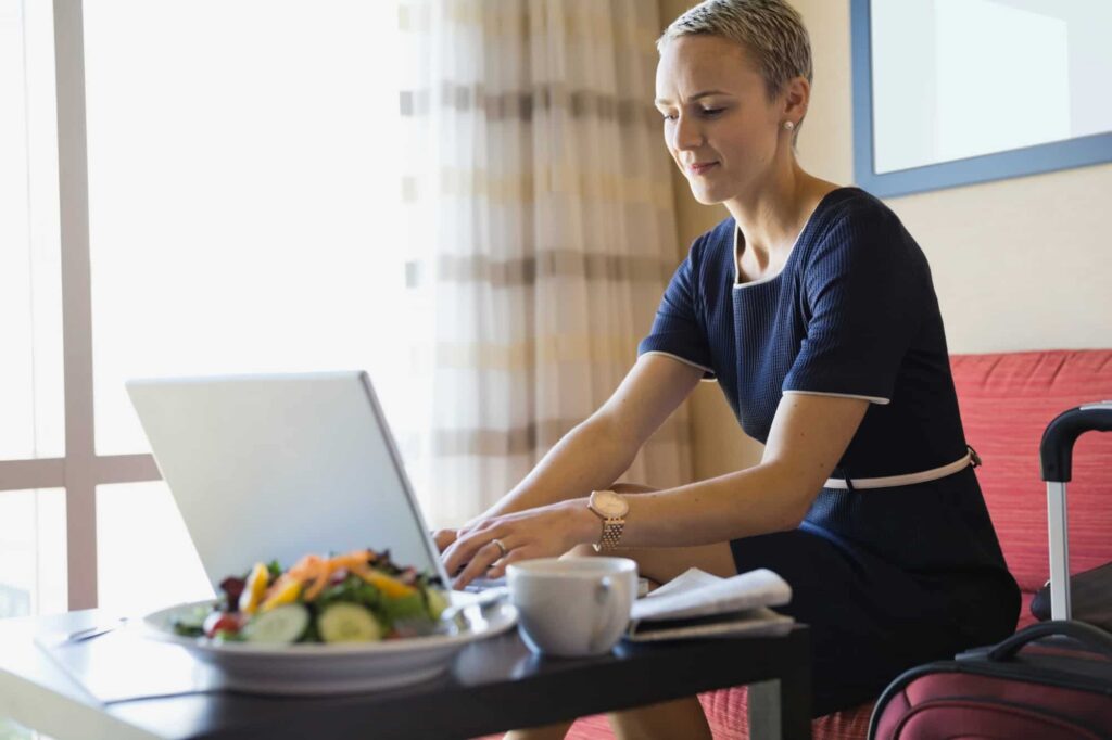Woman working on a laptop in a hotel room, representing remote work or travel use of a VPN, with a focus on privacy and secure internet access outside a trusted home network.