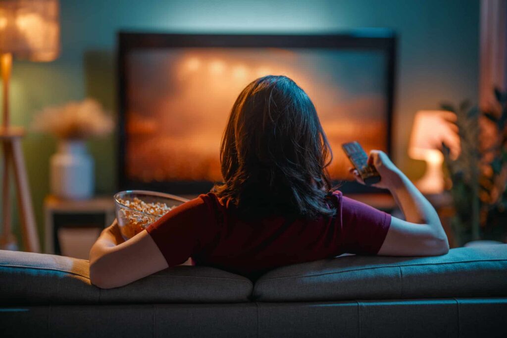 Person sitting on a couch using a laptop and TV remote while watching streaming content, illustrating home internet use, VPN safety, and online privacy.