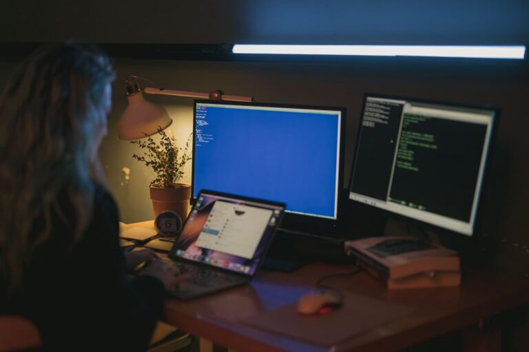 Woman working in a home office using multiple computer monitors, focused on cybersecurity-related tasks in a comfortable workspace.